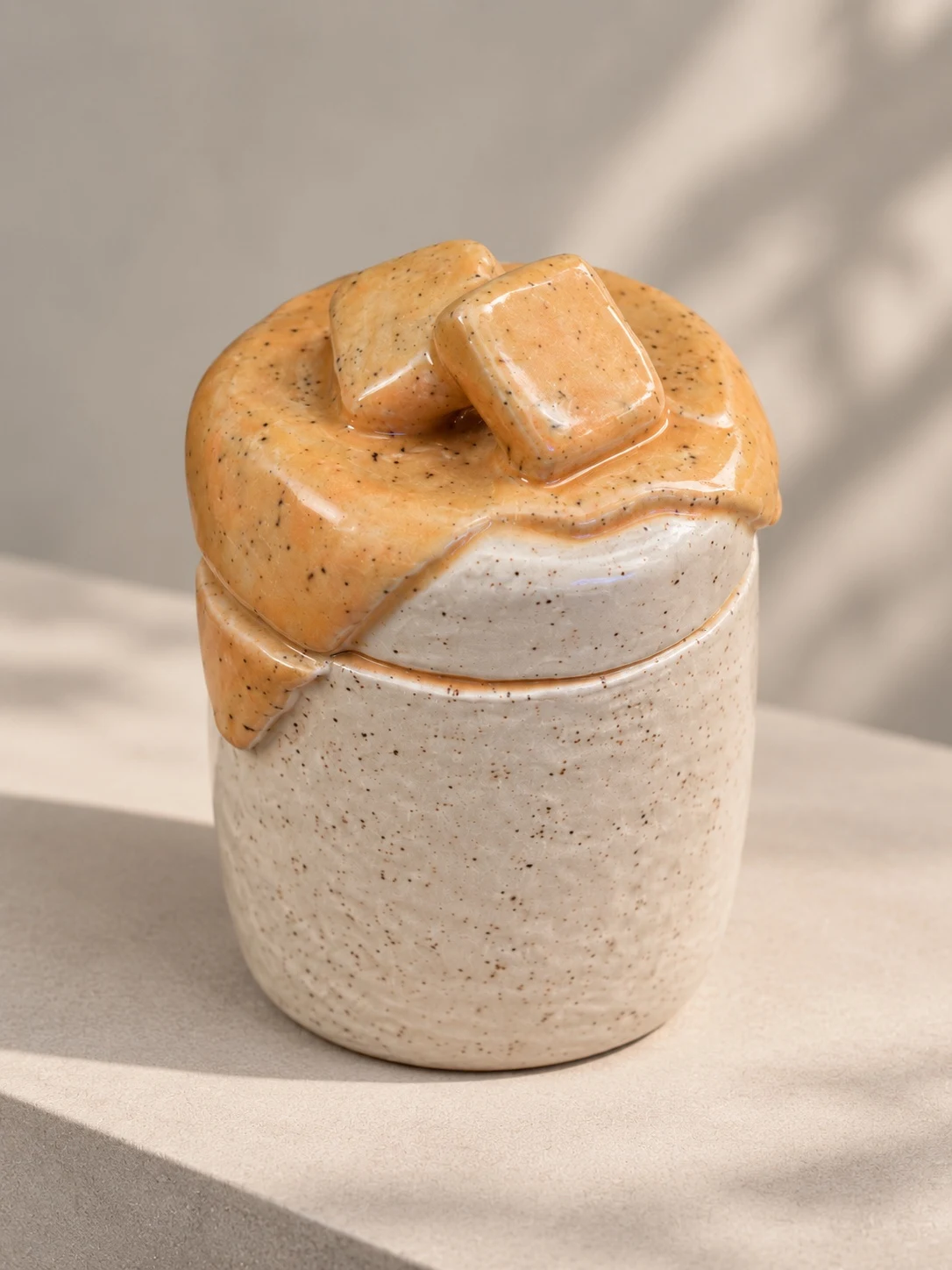 Speckled cream ceramic jar with orange glaze flowing over its lid and two sculpted butter pats